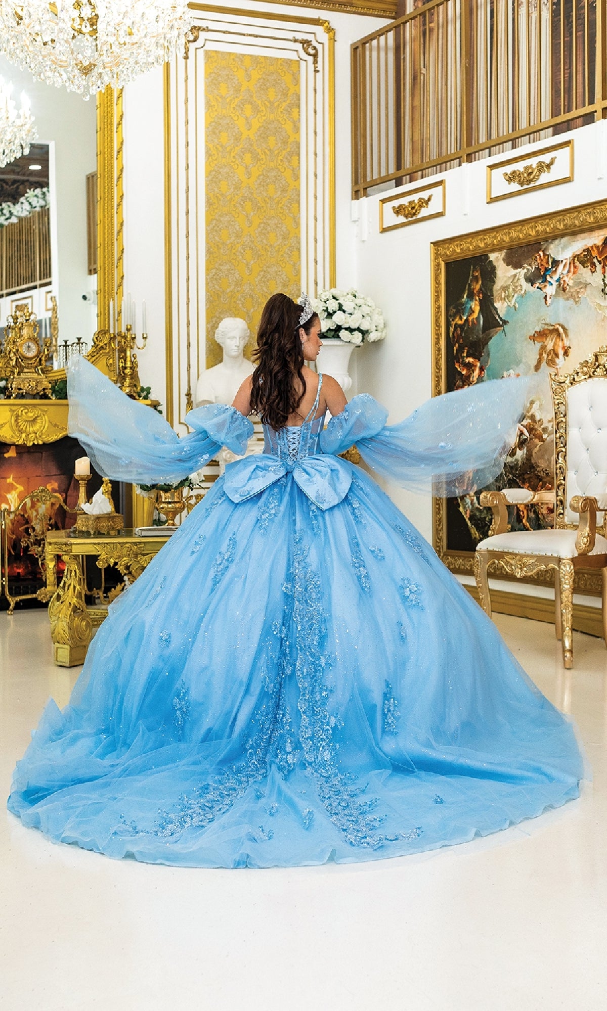 A woman in the Blue Quinceanera Dress 5037 by Dancing Queen, featuring blue glitter and floral details, stands in an ornate gold-accented room with her back to the camera.